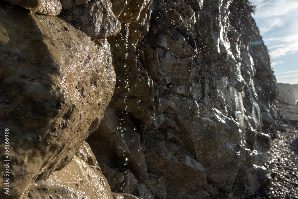 This image offers a detailed close-up of water cascading down a rough, textured rock face on a sunny beach. The sunlight catches individual droplets, highlighting their clarity and the intricate paths