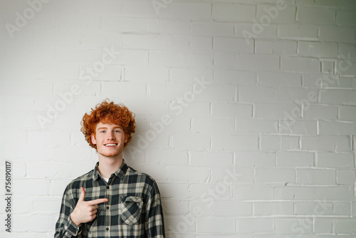 jeune homme souriant roux aux cheveux bouclés et portant une chemise bucheron verte, qui pointe du doigt vers l'espace négatif du fond, copyspace, fond mur briques blanches photo