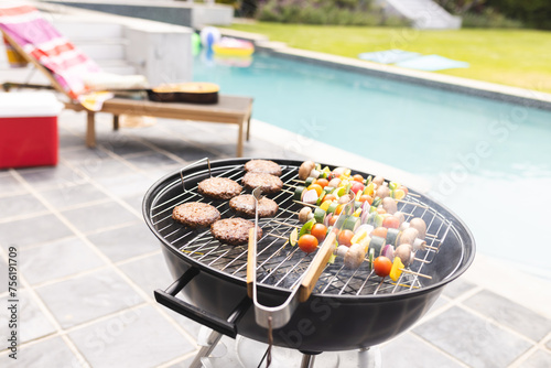 A barbecue grill is loaded with burgers and vegetable skewers, with a pool in the background