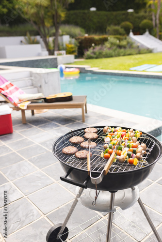 A charcoal grill is cooking burgers and vegetable skewers by a poolside with copy space