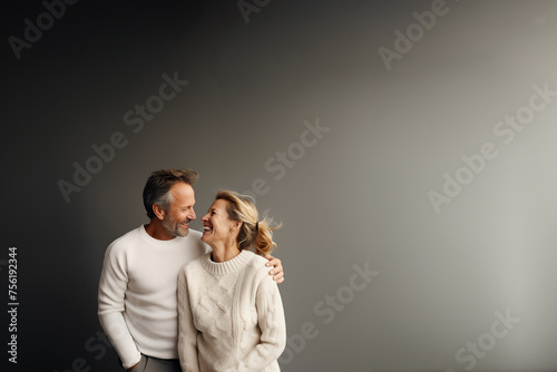 couple hétérosexuel quinqua, 50 ans environ, habillé avec des pulls blancs, qui se tiennent dans les bras et rigolent en se regardant. Fond gris avec espace négatif pour texte copyspace photo