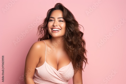 A radiant plus-size female model, age 25, Hispanic, smiling brightly against a gentle pink backdrop, showcasing beauty and positivity.
