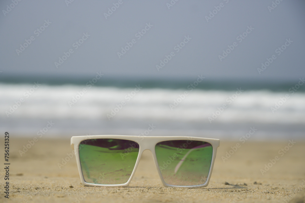 White tinted square wayfarer Sunglass on a beach and waves coming in sea can be seen in the background. Green polarized glasses kept at sand on beach near sea. Sunny day at arabian sea in Goa, India