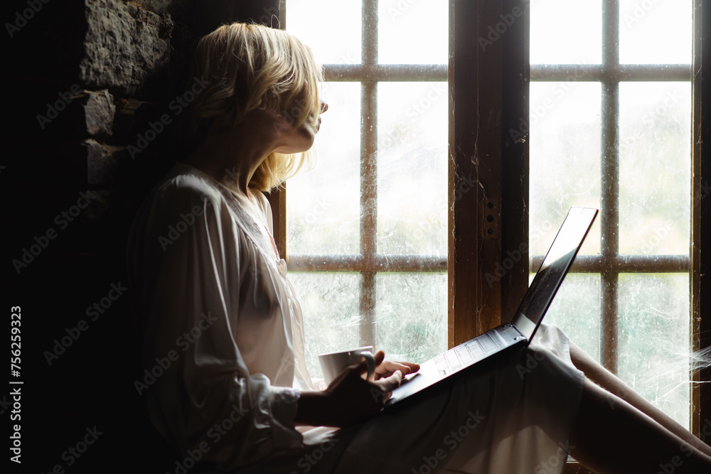 A woman sits with a laptop near the window. A middle-aged woman communicates, works at a laptop. Freelancer, work from home. A woman sits with a laptop near the window. A middle-aged woman communicates, works at a laptop. Freelancer, work from home.