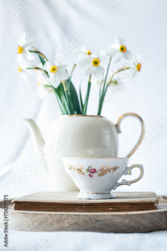 Still life with a blooming bouquet of white daffodils in an elegant porcelain teapot, a baroque porcelain coffee cup and an old book on a textured white background © Jess_Ivanova
