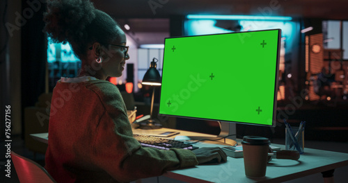 Over the Shoulder: African Female Specialist Working on Computer with Chroma Key Display at Creative Agency. Young Woman Sitting at Her Desk Using Desktop Computer with Mock-up Green Screen © Gorodenkoff