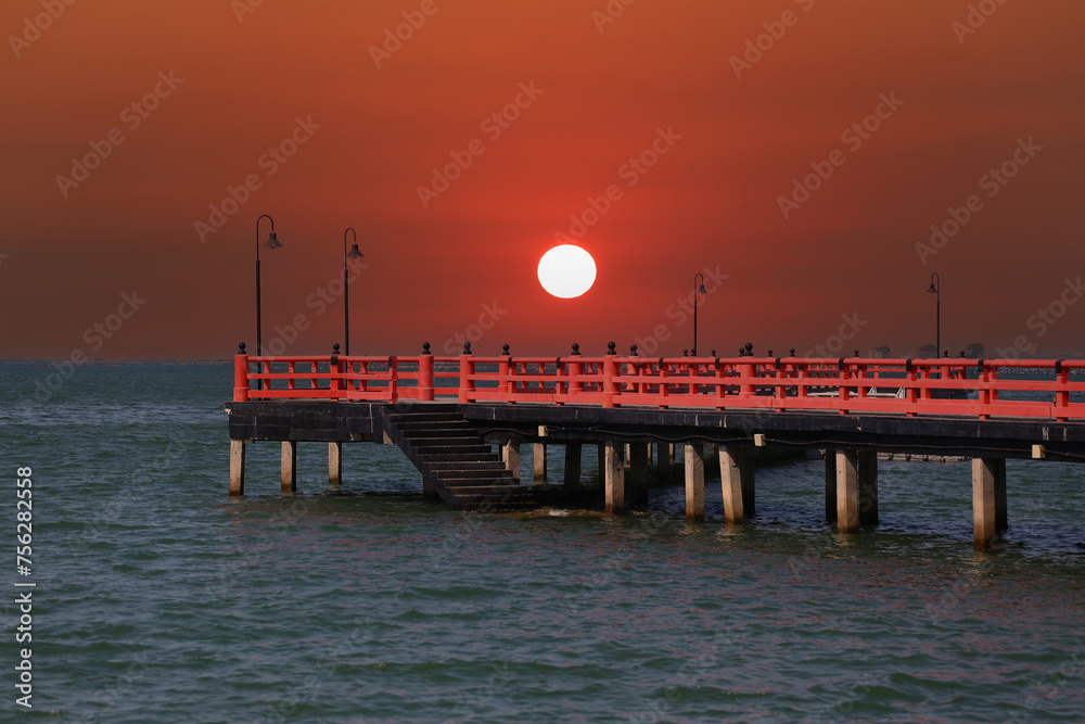 Red bridge that extends into the sea and view of the sunset in the evening time.