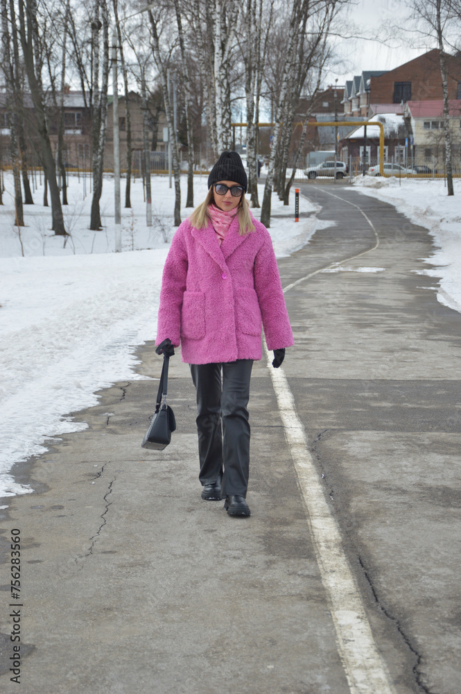 A fashionable Caucasian blonde girl in a bright pink winter fur coat, a black hat and trousers walks along an asphalt road in a winter park.