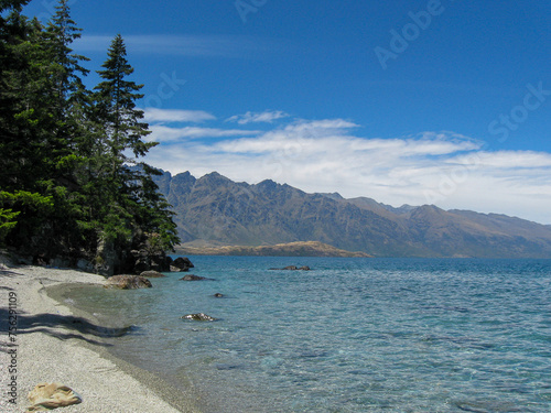 Gravel beach on beautiful, pristine Lake Wakatipu, surrounded by tall green pine trees and with a view of the Remarkables mountain range at Queenstown on New Zealand's South Island