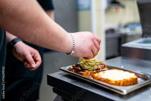 chef adding sliced pickles to a grilled sandwich in a professional kitchen. The sandwich consists of two slices of toasted bread—one topped with grilled meat and the other with a spread.