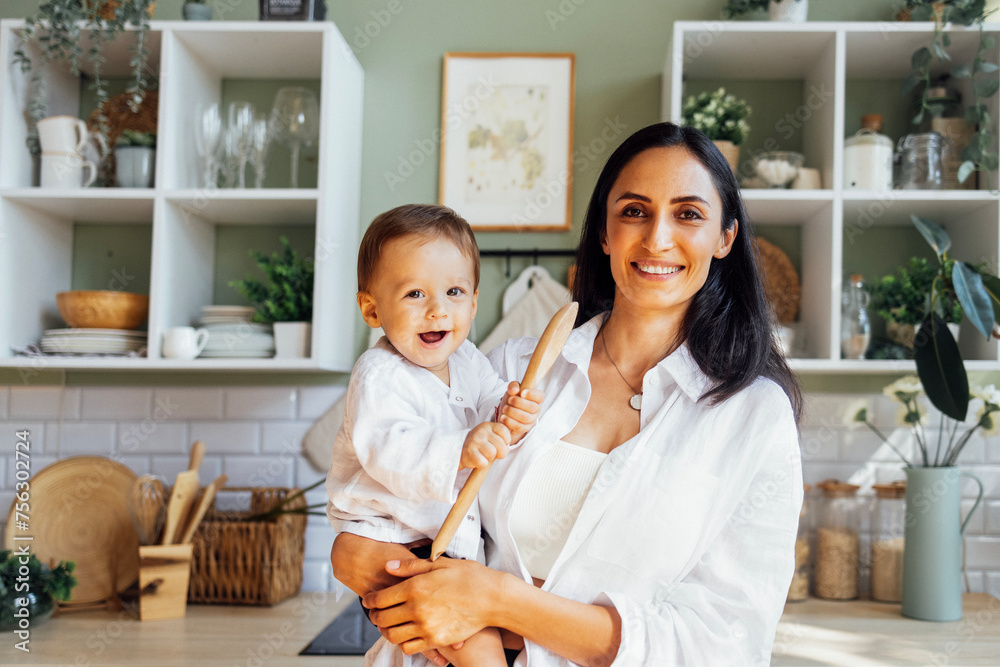 A young mother is cutting greens on a wooden stand in a modern kitchen and her little baby is playin A young mother is cutting greens on a wooden stand in a modern kitchen and her little baby is playin