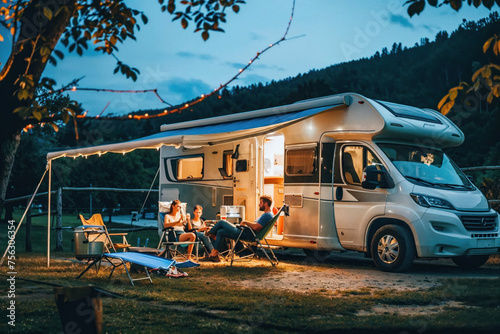 A group of friends unwind and surf the internet on a laptop while sitting by their motorhome at dusk