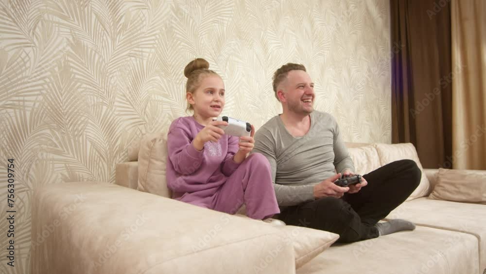 A father spends time with his little daughter, sitting on the sofa and playing a game console. Weekend activities, free time, home entertainment and video games concept. A father spends time with his little daughter, sitting on the sofa and playing a game console. Weekend activities, free time, home entertainment and video games concept.