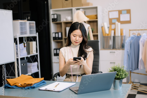  Asian young woman on desk in office of fashion designer and holds tablet, laptop and smartphone on desk.
