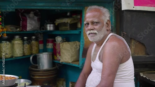 A local aged or old bearded male street food vendor or snack seller is sitting at a roadside shop or stall looking or facing the camera 