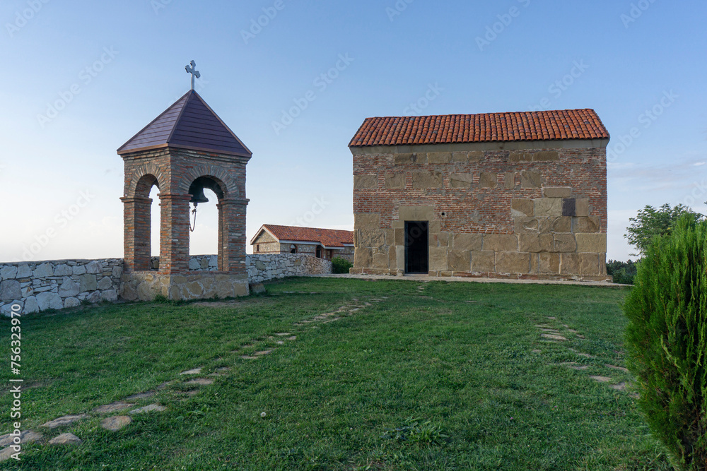 St. Giorgi church of Udzo monastery and bell tower. Tile orange roof, brick and stone wall. Brick arches of the bell tower. Grass yard, blue clear sky.