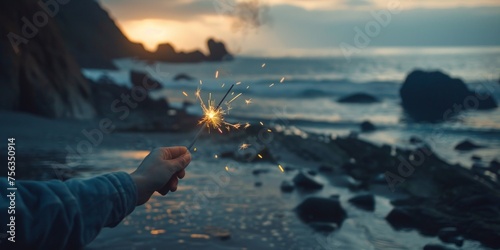 Hand holding a lit sparkler at dusk on a rocky shoreline. © tashechka
