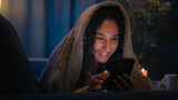 Hispanic woman girl using smartphone under blanket on bed couch reading social media funny network laugh fun laughing enjoy humorous mobile phone illuminated by bright monitor at night gadget addict