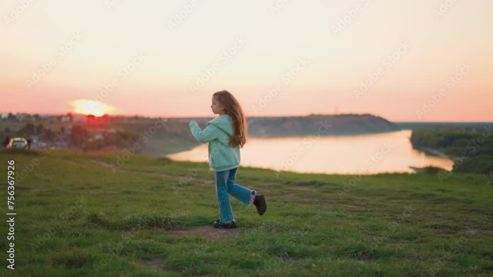 Happy kid runs around on hilly riverbank at sunset. Pretty preschooler girl plays at quiet countryside in evening. Little child enjoys rural vacation Happy kid runs around on hilly riverbank at sunset. Pretty preschooler girl plays at quiet countryside in evening. Little child enjoys rural vacation