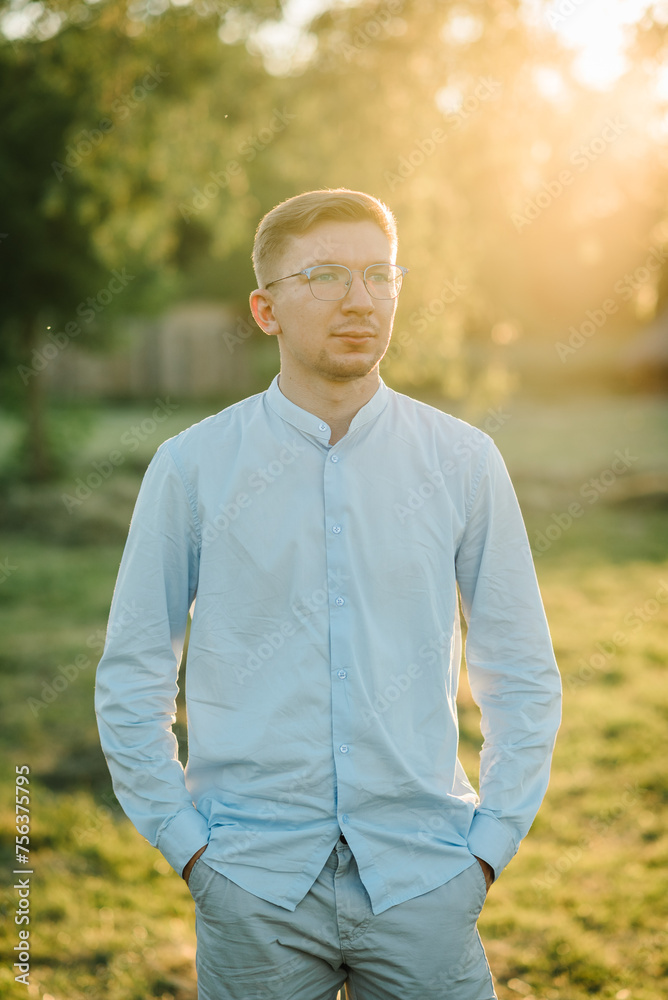 A handsome man in glasses stands on green grass at sunset. Serious male on the field in the sunset background. Male portrait on summer day. A handsome man in glasses stands on green grass at sunset. Serious male on the field in the sunset background. Male portrait on summer day.