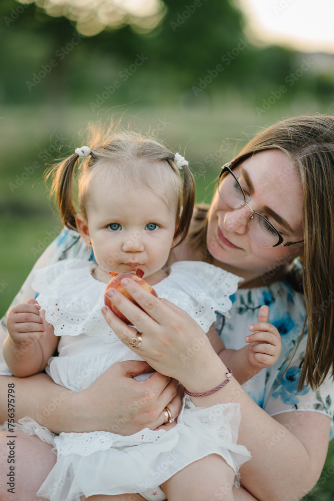 Fototapeta premium Mom feeds a child a peach on a picnic. Mom and girl playing in park at sunset. Mommy and kid spend time together. Portrait of a daughter toddler hugging mother in a field on summer day. Closeup.
