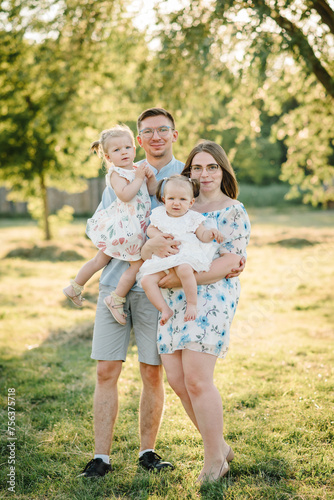 Children hugging parents in nature at sunset. Mom, dad and childs girl walking in the green grass. Happy young family in park spending time together, on vacation, outdoors. Concept of family holiday.