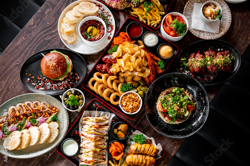 Wallpaper Mural Assortment of delicious meals on a dark wooden table, including a glossy bun burger, onion rings, potato wedges, grilled sandwiches, and a bowl of garnished hummus Torontodigital.ca