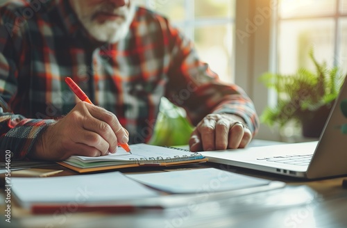 Close up hannd of man writing in notebook with pen while sitting at a wooden desk near a laptop computer, photo