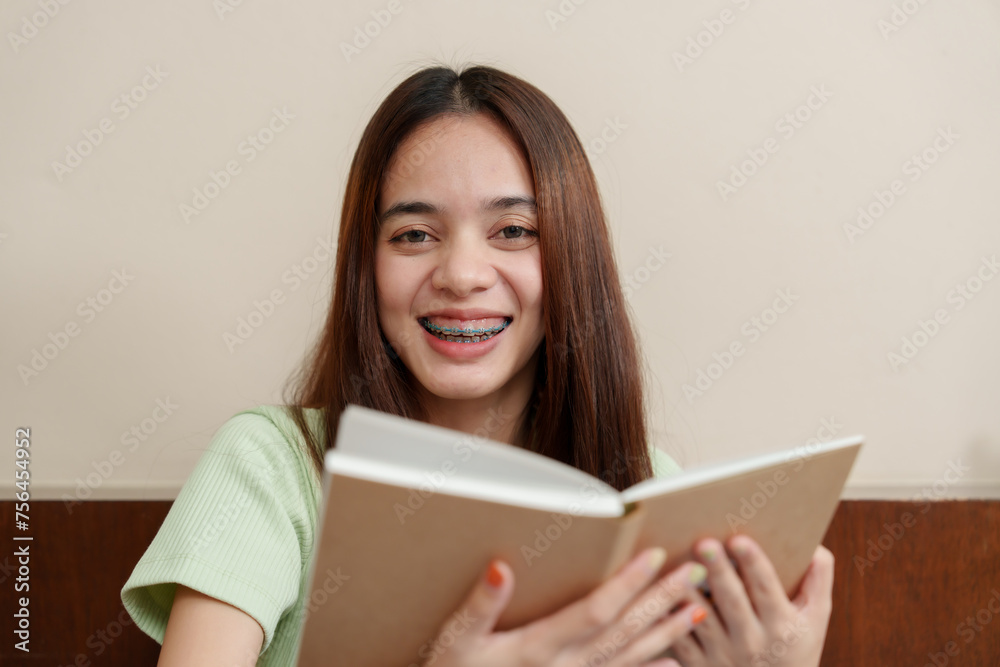 Asian woman in green, holding open book with delight, showcasing braces, in homely setting, joy evident. Gleeful female with dental braces grins holding literature, in cozy corner Asian woman in green, holding open book with delight, showcasing braces, in homely setting, joy evident. Gleeful female with dental braces grins holding literature, in cozy corner