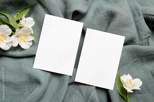 Two empty white cards displayed on a grey linen fabric, accompanied by white alstroemeria flowers with green leaves