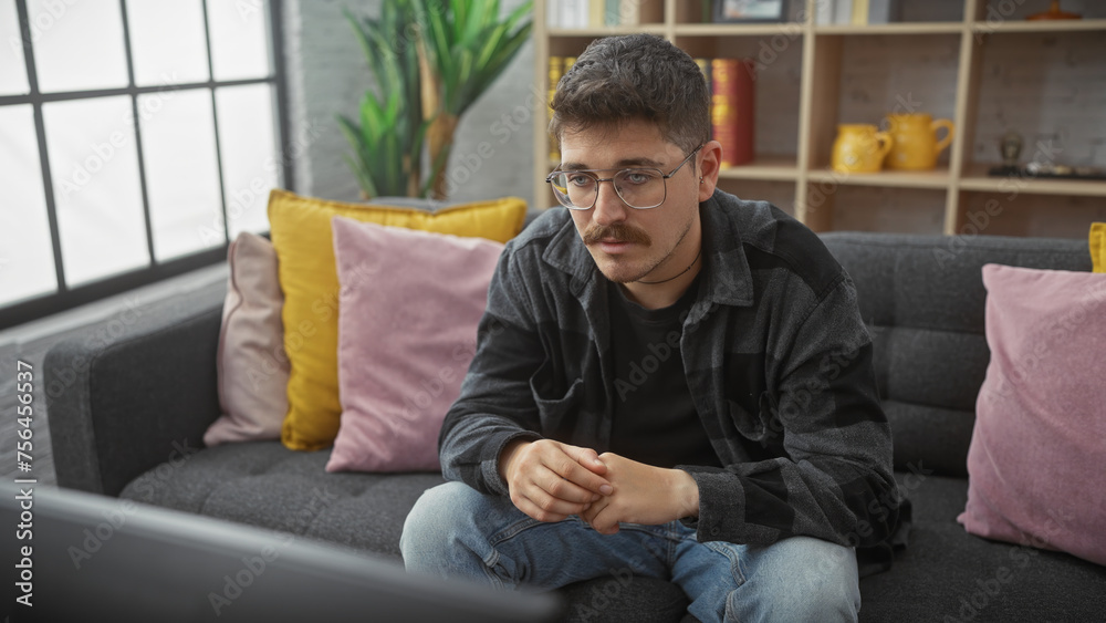 Young hispanic man with moustache in casual clothing sitting thoughtfully on sofa at home, living room interior visible.