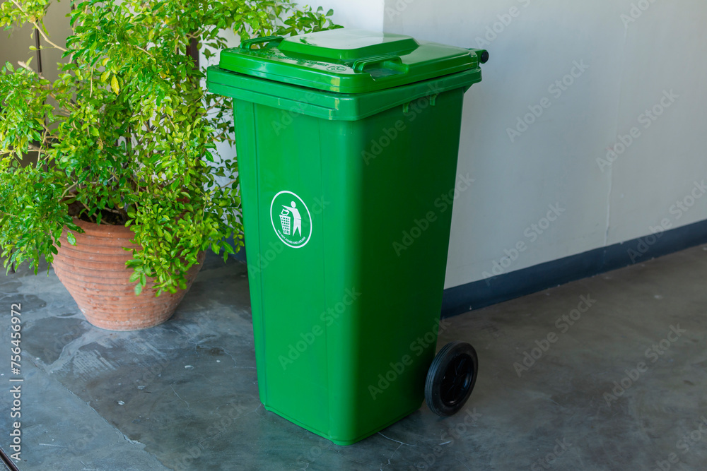 A green plastic wheelie trash bin with a recycling symbol, placed beside a plant pot outside a building. A green plastic wheelie trash bin with a recycling symbol, placed beside a plant pot outside a building.