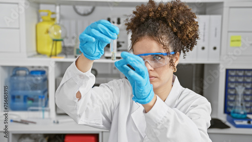A focused hispanic woman with curly hair analyzes a test tube in a laboratory setting  highlighting the importance of scientific research.