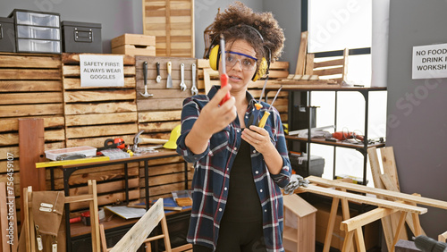 Hispanic woman with curly hair wearing safety goggles in a carpentry workshop holding tools