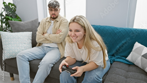 A woman plays a video game while a man watches sullenly in a bright living room, highlighting leisure, gender dynamics, and home life. photo