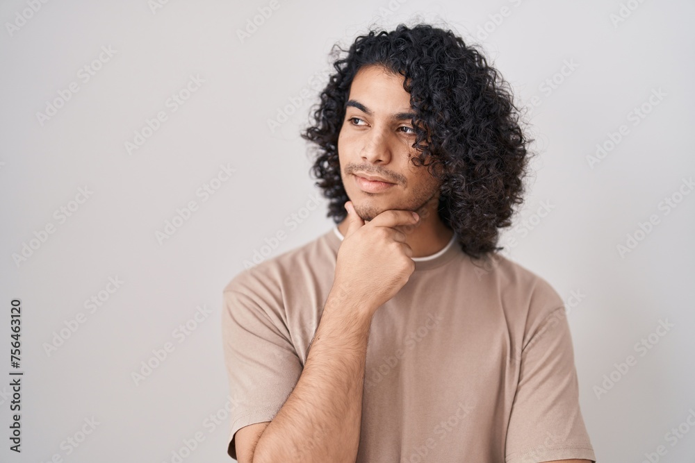 Hispanic man with curly hair standing over white background looking confident at the camera smiling with crossed arms and hand raised on chin. thinking positive.