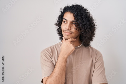 Hispanic man with curly hair standing over white background looking confident at the camera smiling with crossed arms and hand raised on chin. thinking positive. © Krakenimages.com