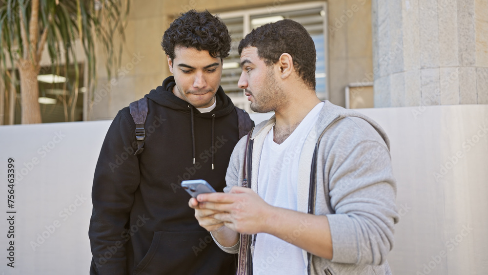 Two hispanic men friends sharing a phone screen together on a sunny city street. Two hispanic men friends sharing a phone screen together on a sunny city street.