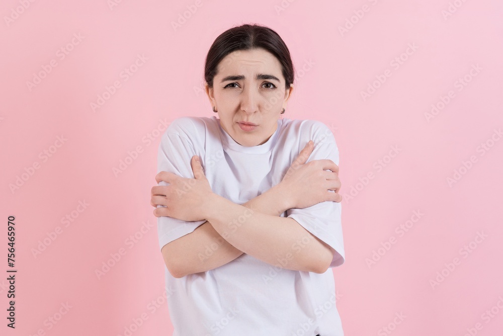 Attractive Caucasian young brunette woman in casual white t-shirt is freezing, she is cold and hugging herself isolated on pink studio background. Attractive Caucasian young brunette woman in casual white t-shirt is freezing, she is cold and hugging herself isolated on pink studio background.