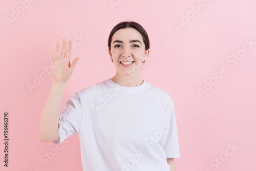 Attractive Caucasian young brunette woman in casual white t-shirt greets, waves hand and looks at the camera isolated on a pink studio background. Says hello.