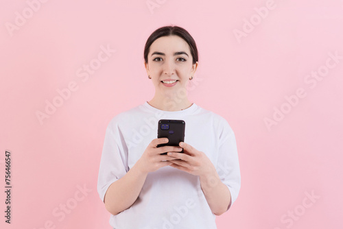 Attractive Caucasian young brunette woman in casual clothes uses mobile phone isolated on pink studio background