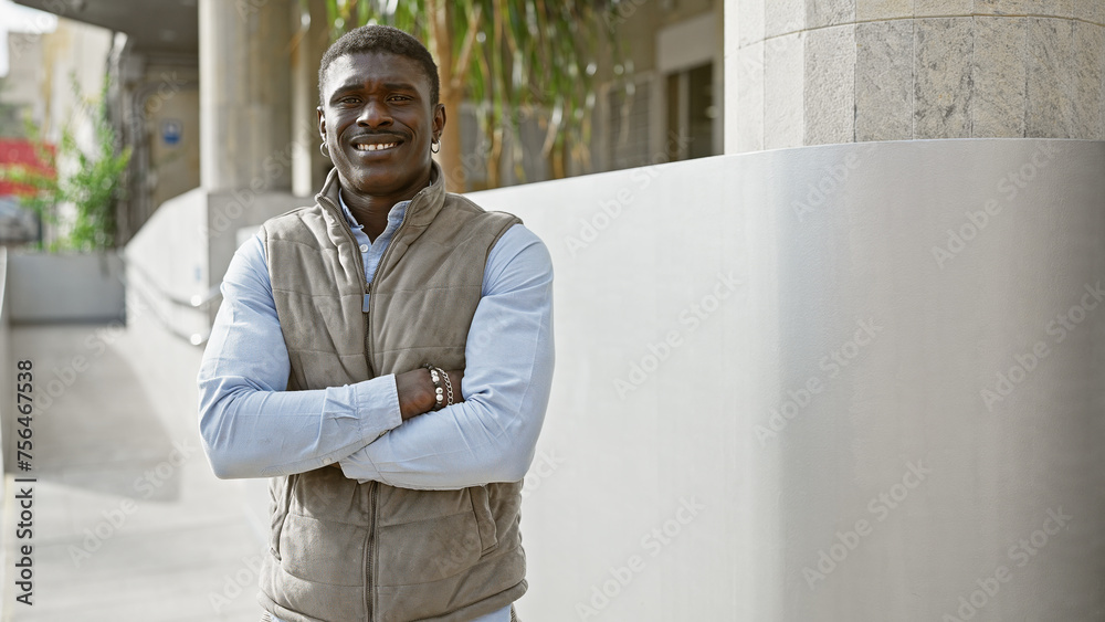 Fototapeta premium Handsome black man with crossed arms standing outdoors in a city environment, smiling confidently.