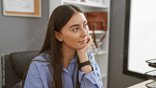 A contemplative young hispanic woman in a striped shirt poses in a modern office setting  exuding professionalism and calm.