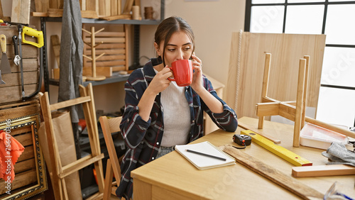 A young hispanic woman enjoys a coffee break in a woodworking workshop surrounded by tools and wood pieces.