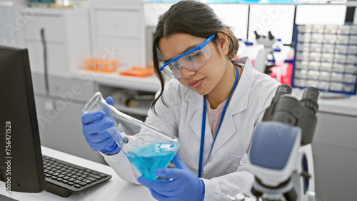 A young hispanic woman scientist analyzing a blue liquid in a flask inside a modern laboratory.