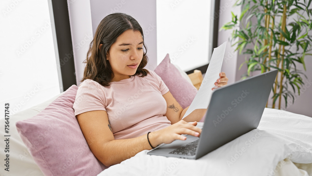 Hispanic woman working on laptop in bedroom with papers and plant in background.