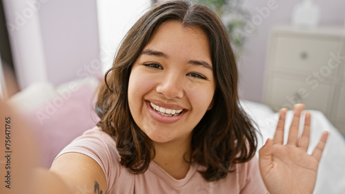 A cheerful young hispanic woman takes a selfie in a cozy bedroom, radiating warmth and positivity.