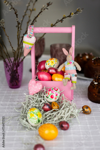 Holiday laid table for Easter celebration with colorful hand-painted Easter eggs in pink wooden basket, toy bunnies, willow branches and Easter cakes