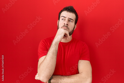 Young hispanic man wearing casual red t shirt with hand on chin thinking about question, pensive expression. smiling with thoughtful face. doubt concept.