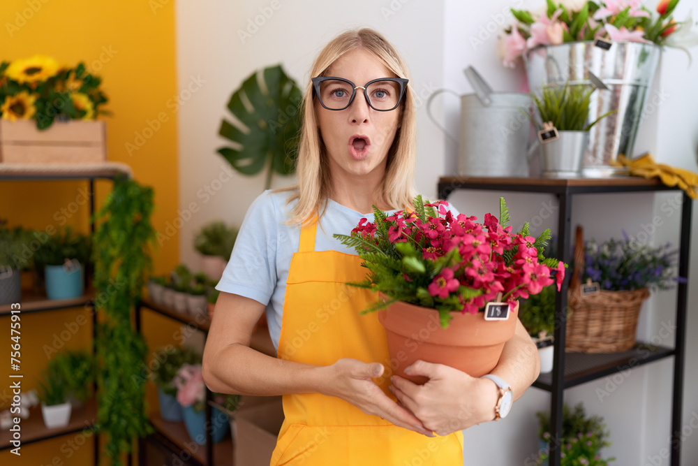 Young caucasian woman working at florist shop holding plant scared and amazed with open mouth for surprise, disbelief face